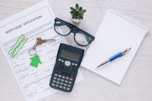 A top-view of a mortgage application marked "APPROVED" on a wooden desk, accompanied by a calculator, a pair of glasses, house keys with a green keychain, and a blank notebook with a blue pen.
