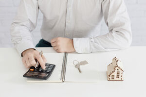 A man in a light gray striped shirt uses a calculator at a desk, with an open notebook, a small wooden house model, and keys beside him, against a white brick wall background.