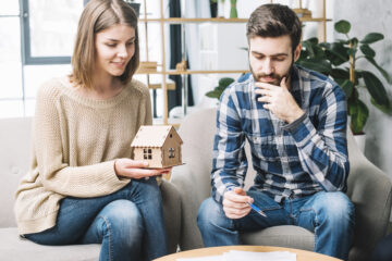 A young couple discussing home ownership while holding a small model house, symbolizing decisions around buying a home or mortgage planning.