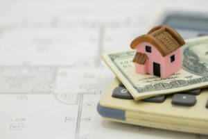 A pink toy house sits on top of a stack of money and a calculator, with a partially visible architectural blueprint in the background. This image suggests a focus on real estate or financial planning.
