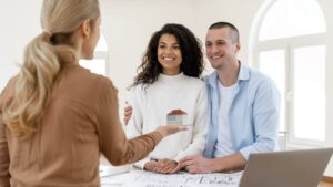 A couple smiles as a real estate agent presents a miniature house model to them, with architectural plans and a laptop visible on the table.